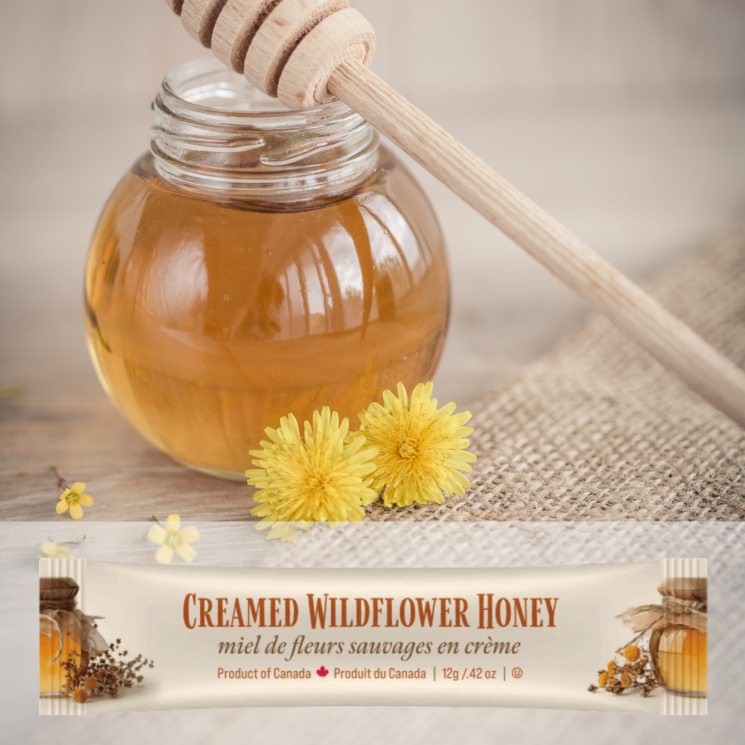 Jar of honey with a wooden dipper, two yellow flowers, and a mini squeeze pack of Tu-Bees  creamed wildflower honey on a textured surface.