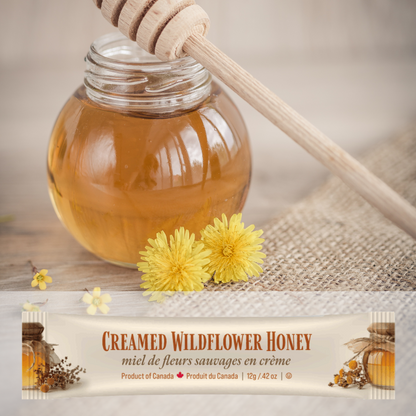 Jar of honey with a wooden dipper, two yellow flowers, and a mini squeeze pack of Tu-Bees  creamed wildflower honey on a textured surface.